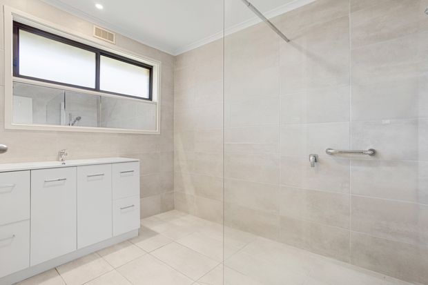 Bright contemporary bathroom with a large frameless glass walk-in shower, full-height pale beige wall tiling, matching floor tiles, a white built-in vanity with drawers and cupboards, and a wide horizontal window with dark trim. Chrome grab rails and tapware provide practical detailing.