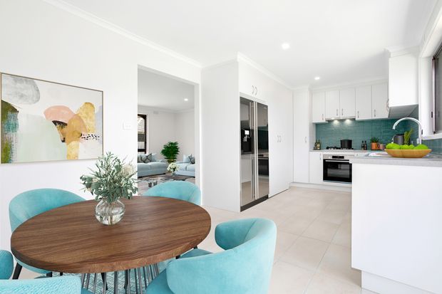 Bright open-plan dining and kitchen interior with a round dark timber dining table and turquoise upholstered chairs in the foreground. A white kitchen with integrated stainless-steel appliances, white cabinetry, pale tiled flooring, and a teal subway-tile splashback occupies the right side, while a living room is visible through the opening beyond.