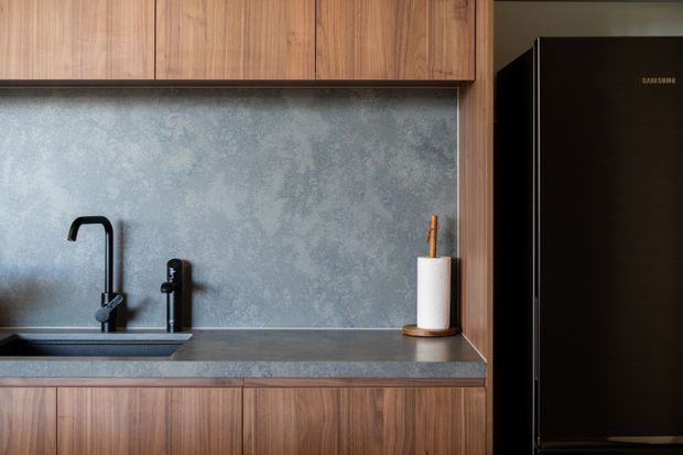 Close-up view of a contemporary kitchen featuring warm timber cabinetry, a mottled grey backsplash and benchtop, a black sink and tapware, and a tall black refrigerator.