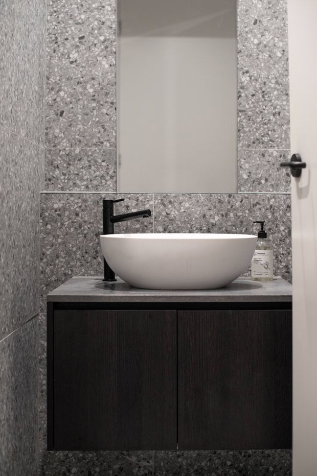 Compact contemporary bathroom vanity featuring a white vessel basin, matte black wall-mounted tapware and soap dispenser, dark timber cabinetry, and grey terrazzo-look tiled walls and benchtop. A large frameless mirror sits above the vanity.