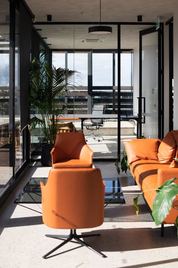 A bright contemporary office lounge with floor-to-ceiling glass partitions and extensive glazing. The space contains vivid orange leather lounge chairs and a matching sofa, a glass coffee table, indoor palms, black-framed doors and partitions, and a workstation visible beyond the glazed divider. Strong sunlight creates sharp shadows across the light terrazzo or polished concrete floor.
