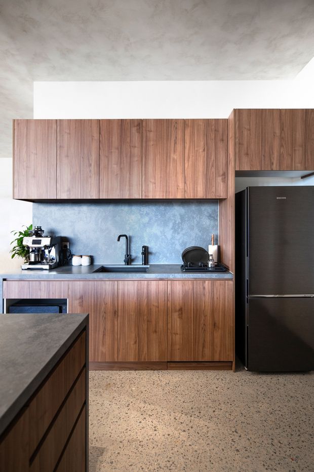 A contemporary kitchen with dark walnut-toned timber cabinetry, a blue-grey textured splashback, black fixtures and appliances, and a terrazzo-style speckled floor. The compact space includes a partially visible island, integrated cabinetry, sink, coffee station, dish rack, and tall refrigerator. A mottled plaster-like ceiling adds texture.