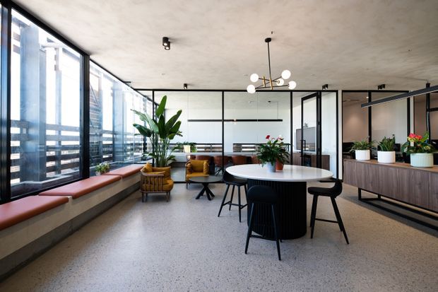 A bright contemporary open-plan interior with a dining and breakout seating area, full-height black-framed glazing, exposed concrete-style surfaces, terrazzo flooring, indoor plants, and a built-in window bench. A timber-toned kitchenette or service counter occupies the right side, while glass partitions and pendant lighting define the workspace-like setting.
