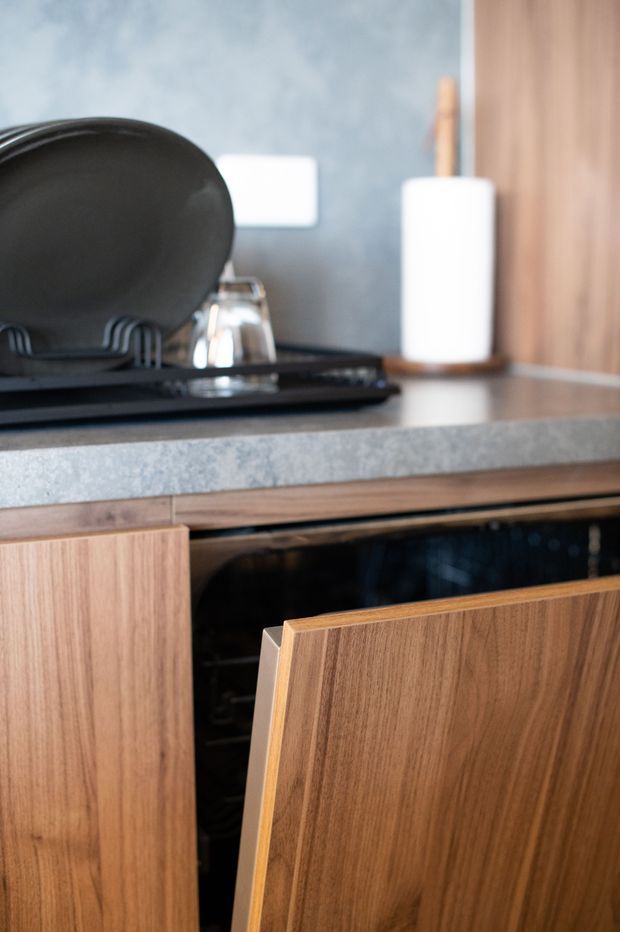 Close-up of a contemporary kitchen or kitchenette showing a dark stone-look benchtop, warm timber cabinetry with an open dishwasher, black dish rack, glassware, and a white paper towel holder. The cabinetry has clean flat-panel fronts and the wall behind is finished in a mottled blue-grey surface.