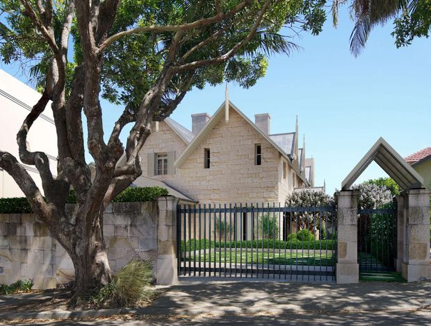 A street-facing view of a substantial light-stone residential building behind a masonry boundary wall and dark metal security gates. The house has prominent steep gables, pitched roof sections, projecting chimneys, and small multi-pane windows. Mature trees and carefully maintained planting frame the frontage, with a distinctive pointed-gable pedestrian gate at the right.