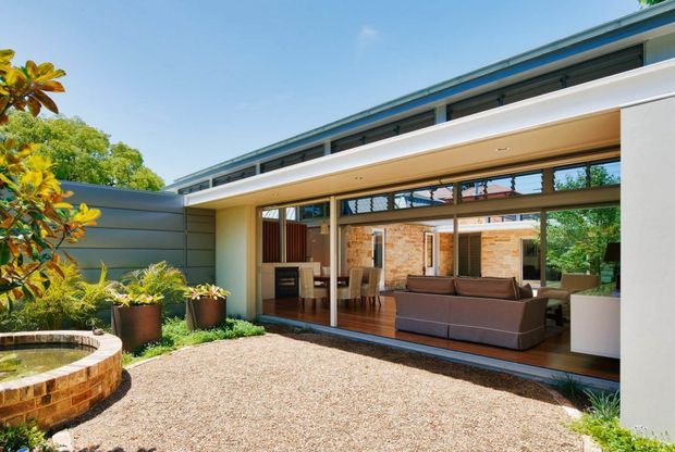 A bright courtyard view of a modern single-storey home, showing a broad covered outdoor living area with timber decking, sliding glass doors, and a visible interior living room and kitchen. The facade combines pale rendered walls, horizontal metal cladding, timber-framed glazing, and exposed brickwork. A curved brick-edged garden feature and planted borders sit within a gravel-paved courtyard.