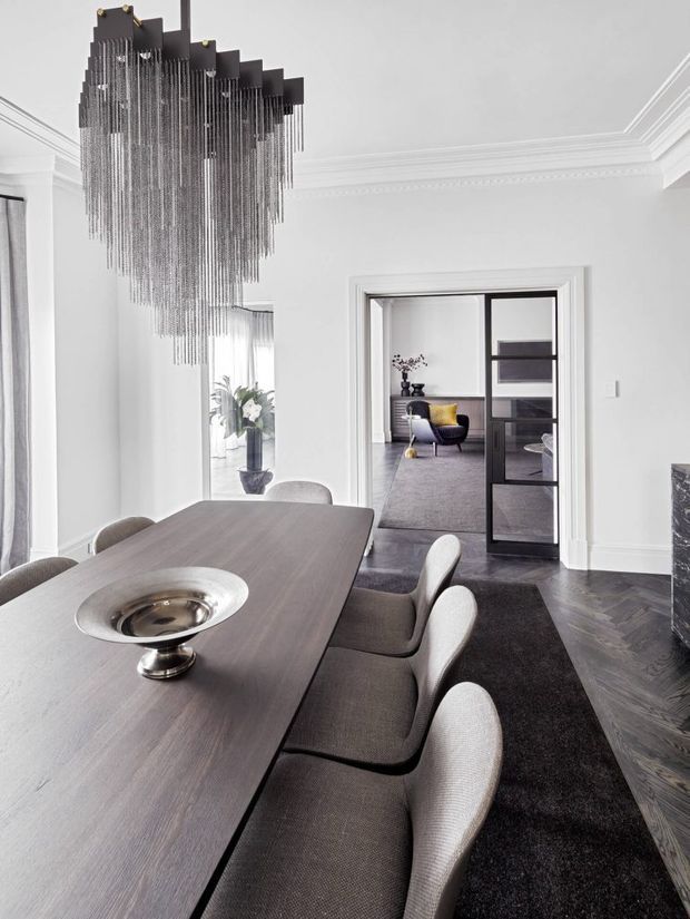 Elegant formal dining room with a long dark timber dining table, upholstered dining chairs, a dramatic tiered black pendant chandelier, white decorative wall mouldings, and dark herringbone flooring. A glazed black-framed doorway opens to a refined adjoining sitting room or lounge.
