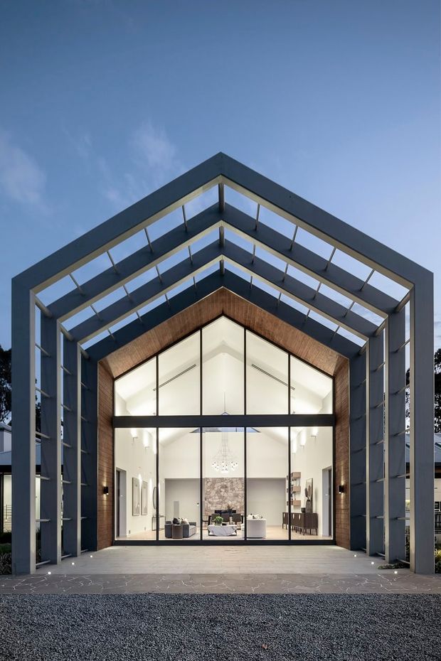 A dramatic dusk exterior view of a contemporary, gable-ended building framed by a large open steel structure. The facade features extensive floor-to-ceiling glazing, dark metal framing, warm timber lining beneath the gable, and a brightly illuminated interior with visible living and dining furnishings. A paved entry terrace and gravel foreground lead to the glass frontage.