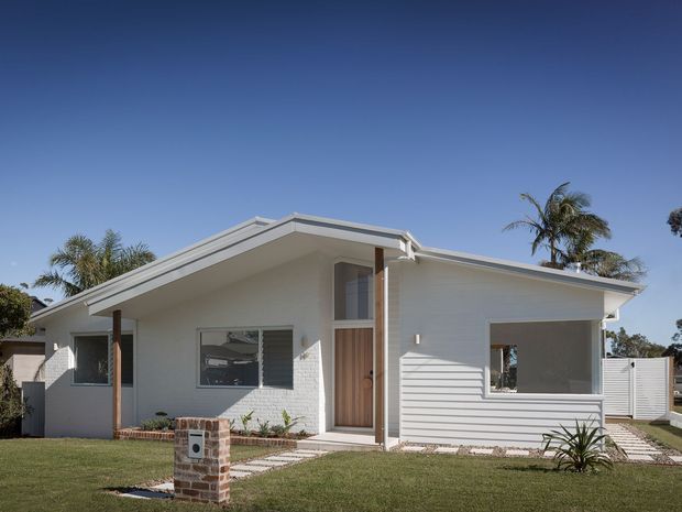 Front exterior view of a low-profile single-storey home with a light-painted horizontal weatherboard facade, broad asymmetrical gable roof, projecting eaves, timber entry door, large windows, landscaped lawn, and a brick-clad utility or mailbox feature in the foreground. Palm trees and clear blue sky surround the property.