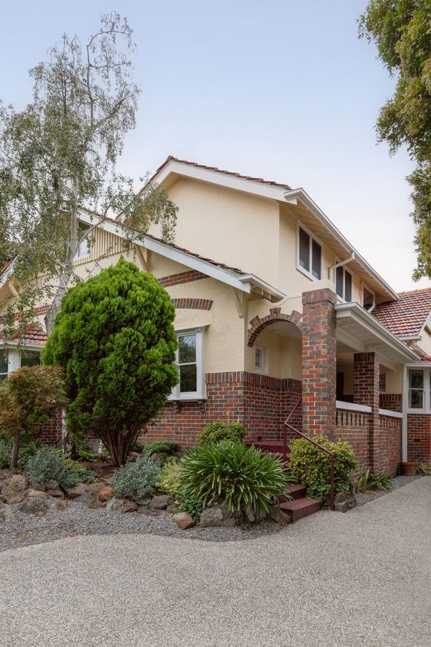 Exterior view of a multi-storey suburban residence with cream stucco walls, red and dark brickwork, tiled pitched roofs, gabled forms, covered porch, arched entry opening, and landscaped front garden with steps and handrail.