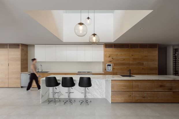 Contemporary open-plan kitchen with a large marble-clad island, integrated white cabinetry, warm timber wall and base cabinetry, three black bar stools, and three pendant lights suspended beneath a dramatic recessed ceiling opening. A wine rack is visible at the far right.