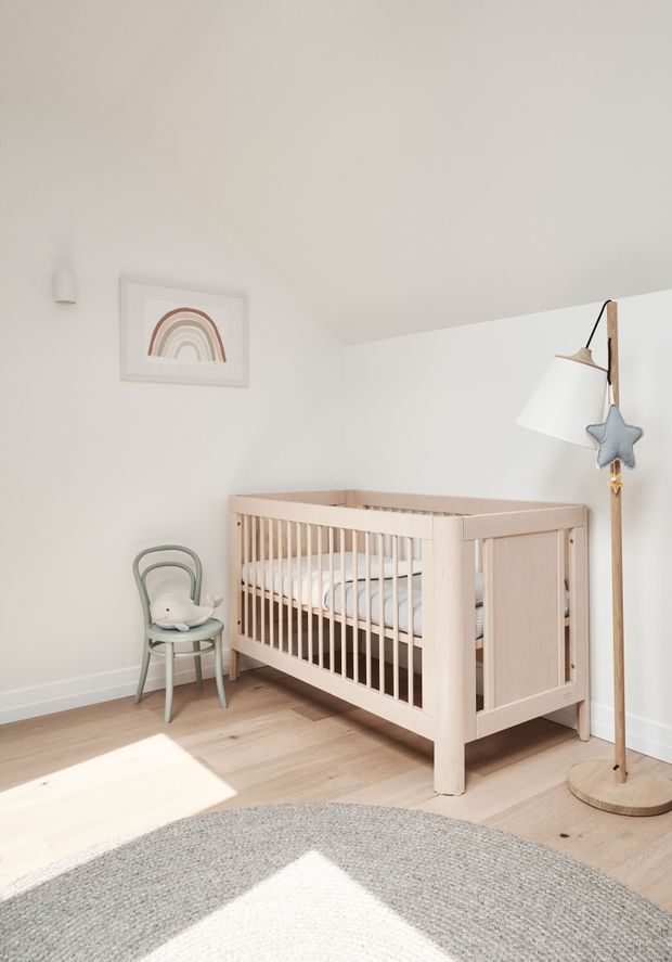 Bright, softly styled nursery with a light timber cot, pale timber flooring, white walls, a small muted rainbow print, child-sized chair, floor lamp, and large neutral round rug. Natural sunlight creates a warm, airy atmosphere.