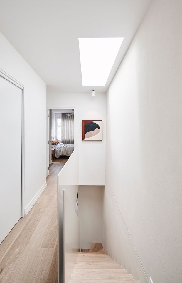 A bright, minimalist residential hallway viewed from the top of a stairwell. White walls and ceiling surround pale timber flooring and matching stairs, with a simple white balustrade and recessed stair opening. A large rectangular skylight illuminates the corridor, while an abstract framed artwork and a glimpse of a bedroom add visual interest. The space has clean contemporary lines and a restrained neutral palette.
