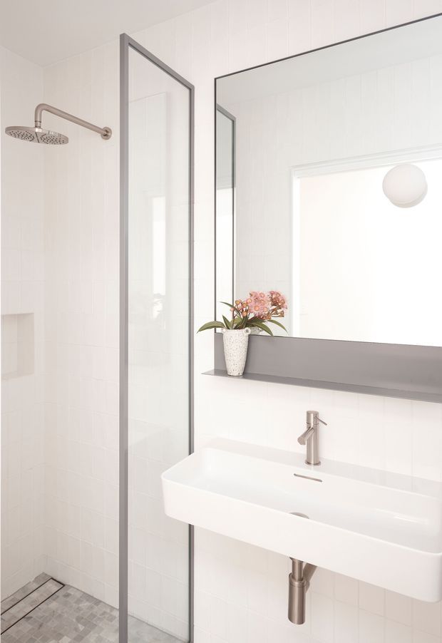 Bright, minimalist bathroom with white tiled walls, a frameless glass shower screen, wall-mounted rainfall shower, grey mosaic shower floor, and a sculptural white basin with brushed metal tap and exposed plumbing. A large mirror and narrow grey shelf sit above the basin, with a small floral arrangement adding colour.