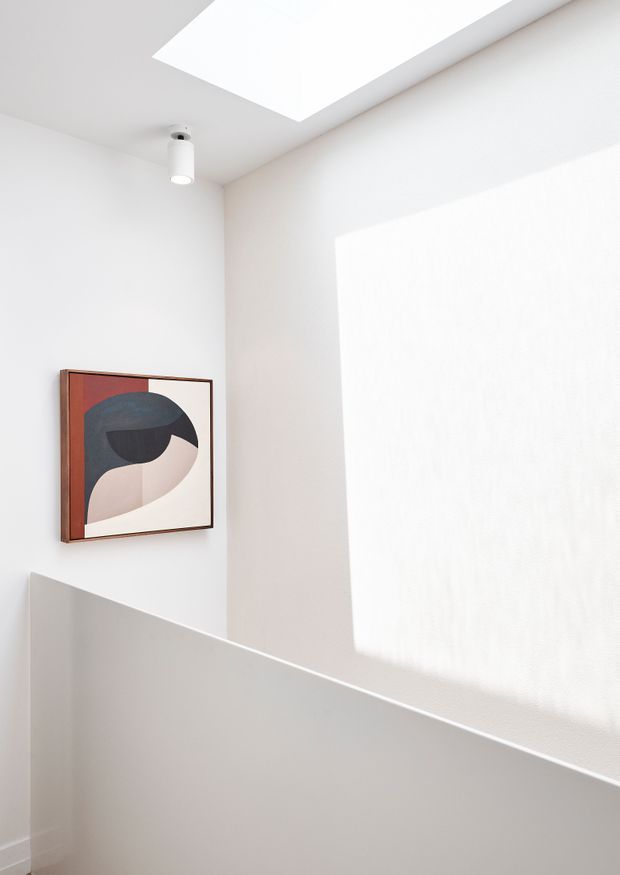A bright minimalist stairwell or landing with white painted walls, a large rectangular skylight, and a simple cylindrical ceiling spotlight. A half-height white stair balustrade crosses the foreground, while an abstract geometric artwork in a slim dark wood frame provides the only strong colour accent. Natural light creates a large rectangular patch of illumination on the wall.