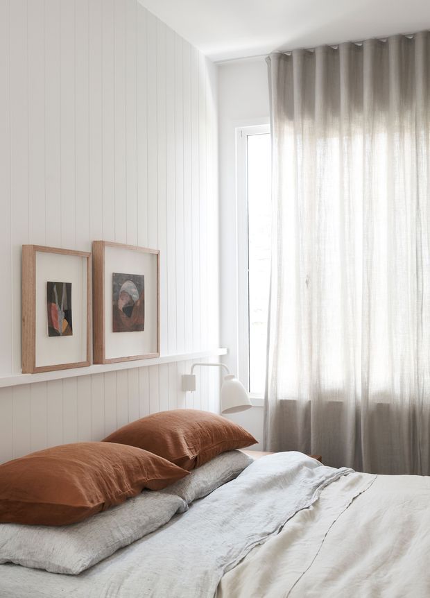 A softly lit bedroom with a white vertical-panelled wall, a narrow floating picture ledge displaying two framed artworks, and a large window dressed with sheer grey curtains. The bed has layered white and light-grey linen bedding with rust-brown pillows, creating a calm, warm and understated interior.