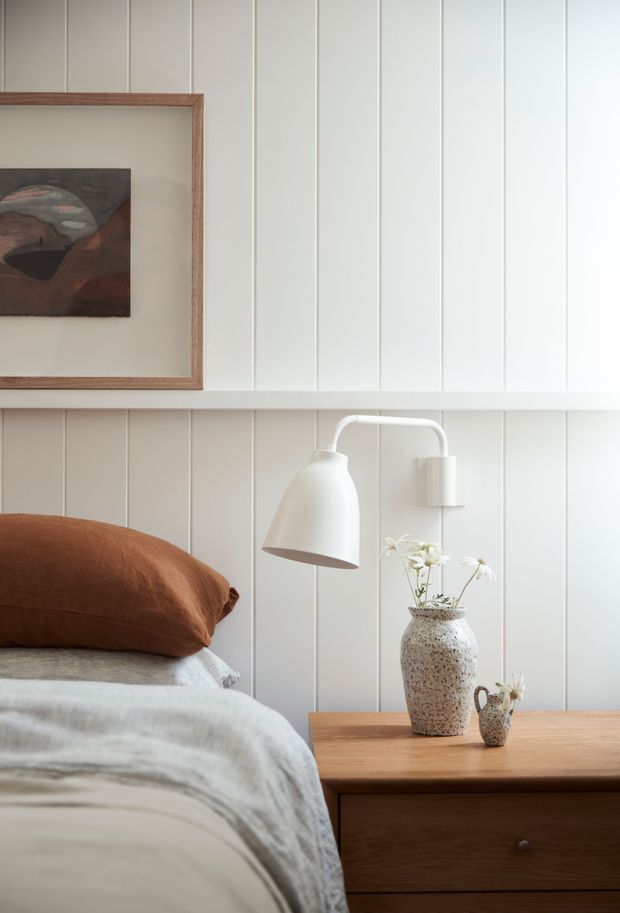 A softly lit bedroom vignette featuring white vertical wall panelling, a timber-framed artwork, a wall-mounted white reading lamp, and a warm timber bedside table. The bed has layered white and pale grey bedding with a rust-brown pillow, complemented by ceramic vases and delicate flowers.