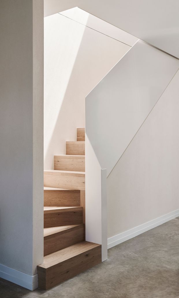 Minimalist interior view of a compact staircase with light timber treads and risers, enclosed by crisp white walls and a sculptural angular white balustrade. Natural light creates strong geometric shadows, while a grey polished concrete floor and simple white skirting complete the restrained modern composition.
