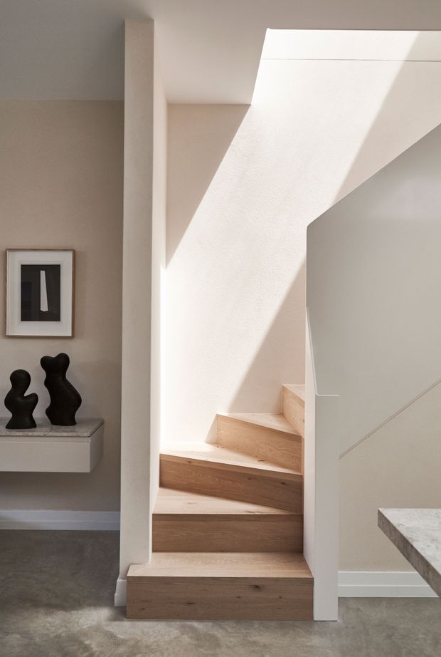 A minimalist interior stairway with pale timber open-riser steps, smooth warm-white plaster walls, and strong sunlight casting geometric shadows. A slim white balustrade and concealed stair structure create a clean sculptural form, while a small floating console with dark abstract sculptures and framed artwork sits beside the stairs. A concrete-like floor and a partial stone benchtop are visible.