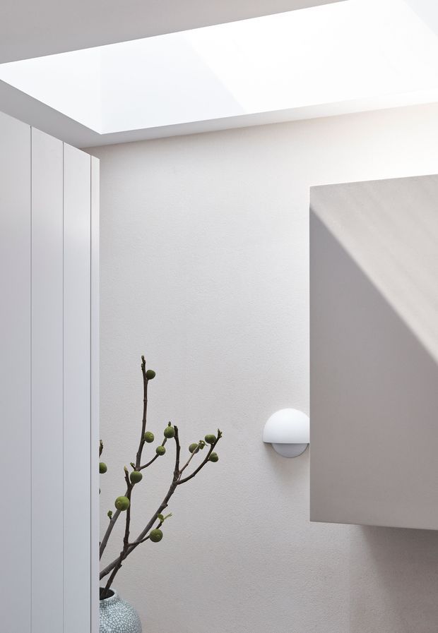 Bright minimalist interior detail with white textured plaster walls, full-height white built-in cabinetry, a large rectangular skylight, a wall-mounted half-dome light, and a sculptural branch in a patterned vase. Strong natural light creates geometric shadows across the walls and ceiling.