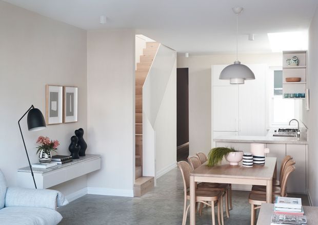 Bright open-plan residential interior showing a dining area and kitchen, with a living room edge in the foreground. The space features pale walls, grey flooring, white cabinetry, a timber dining table with woven chairs, a sculptural pendant light, minimalist console joinery, and a staircase with light timber treads.