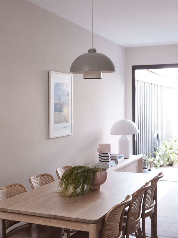Bright, softly styled dining room with a large light timber dining table surrounded by woven cane chairs. A sculptural grey pendant hangs above the table, while a white dome lamp, framed artwork, books, and a plant decorate the sideboard. The room opens to a sunlit outdoor area through large dark-framed glazing.