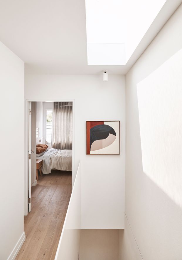 A bright minimalist hallway or landing with white walls, pale timber flooring, a large skylight, and strong sunlight casting geometric shadows. An open doorway reveals a bedroom with a bed and curtains, while an abstract framed artwork adds a muted colour accent.