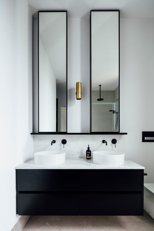 Contemporary bathroom vanity with two round white vessel basins set on a white marble-look benchtop, black floating cabinetry, black wall-mounted tapware, and tall black-framed mirrors. A brass cylindrical wall light adds warmth, while the mirrors reflect a glass shower enclosure with dark fixtures. The space is bright, refined, and high-contrast with clean modern detailing.