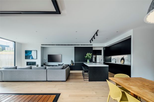 Open-plan contemporary living and kitchen area with a large light-grey sectional sofa, dark built-in cabinetry and media wall, central black kitchen island, pale timber flooring, and a timber dining table with yellow chairs. Floor-to-ceiling glazing and a skylight bring in abundant natural light, creating a clean, spacious interior.
