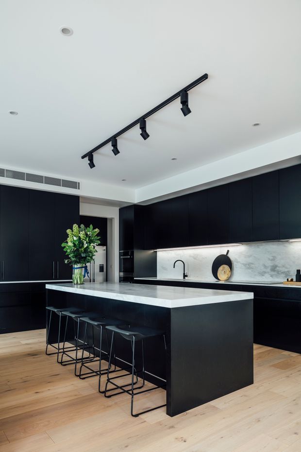 A contemporary kitchen with a large black island, integrated black cabinetry, a pale stone waterfall benchtop, and a light stone backsplash. The island includes seating for four on slim black stools. Track lighting and recessed ceiling lights illuminate the space, while pale timber flooring adds warmth. A sink and black tap are positioned along the rear benchtop, with a vase of greenery on the island.
