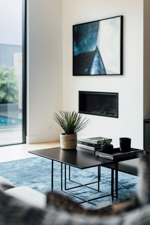 A refined contemporary living room with a black rectangular coffee table, slim black metal legs, a blue-grey area rug, potted plants, stacked books, and a decorative tray. A linear built-in fireplace is set into the white wall beneath abstract artwork. Full-height glazing at the side provides bright natural light and a glimpse of an outdoor pool area.