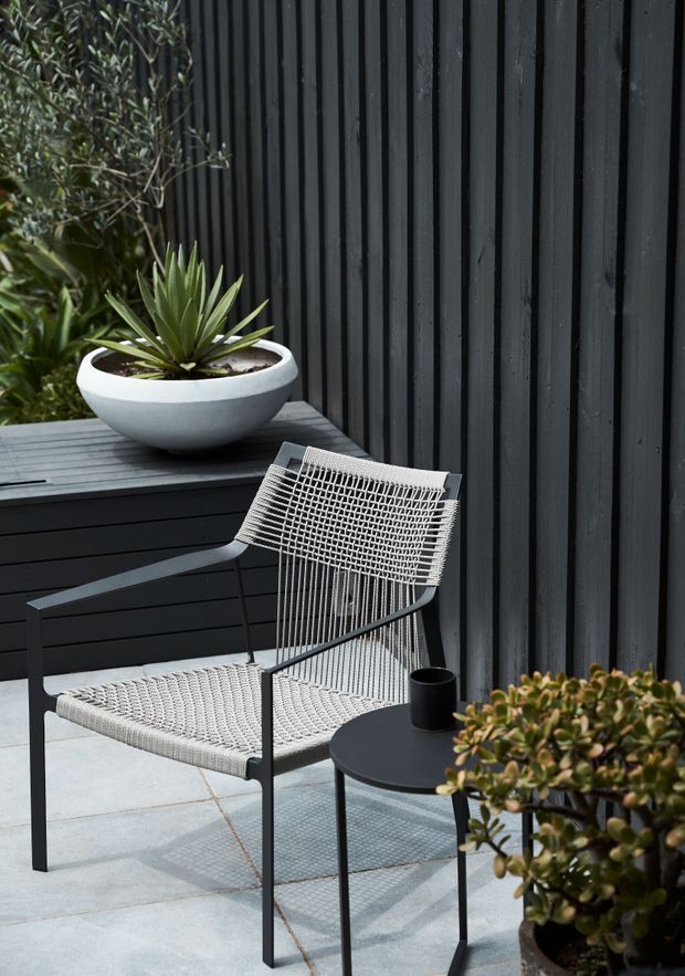 A minimalist outdoor courtyard or terrace with pale square-paved flooring, a black vertical timber-clad wall, and dark built-in planter or bench joinery. A woven sling lounge chair and small round black side table create a simple seating area, accompanied by sculptural potted succulents and greenery.