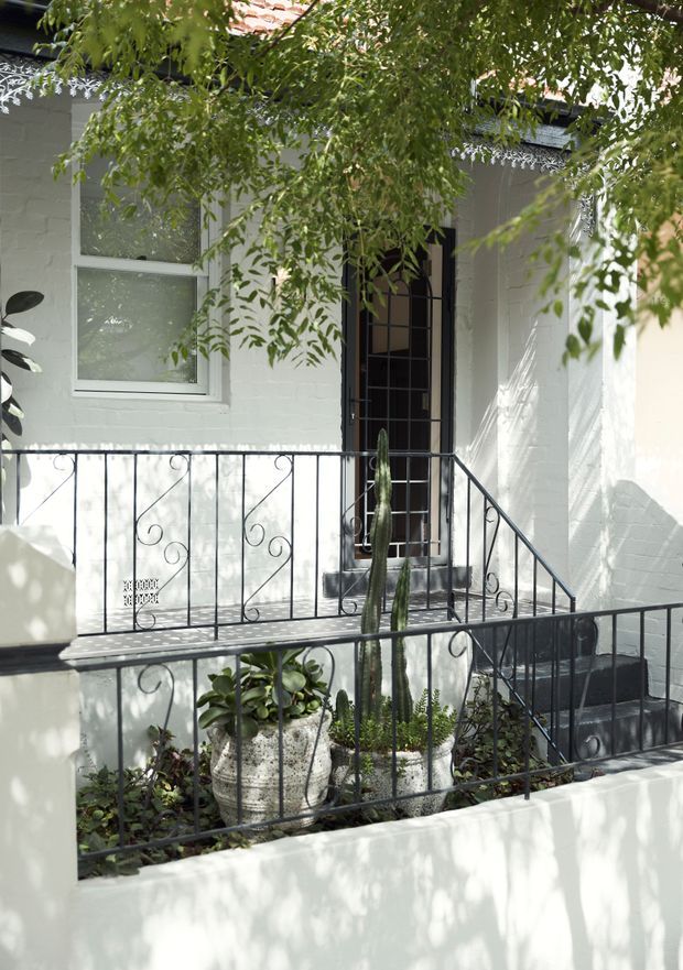Bright exterior view of a small raised front entry porch with white-painted brick walls, a tall black glazed security door, white-framed window, terracotta tiled roof edge, and decorative black wrought-iron railings. Potted succulents and cactus plants occupy the enclosed planter area, with dappled tree shadows across the facade.