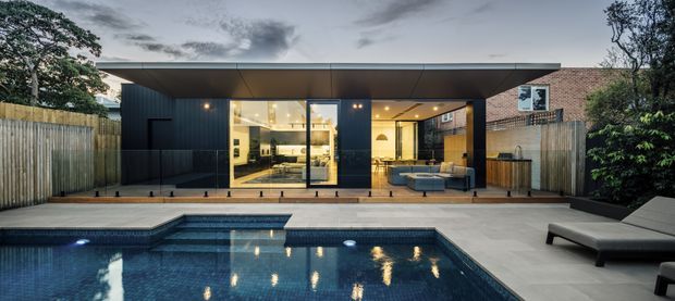 Twilight exterior view of a contemporary single-storey pavilion-style home opening onto a swimming pool. The dark-clad structure has a broad flat roof, extensive floor-to-ceiling glazing, timber screening and a sheltered outdoor seating area. A paved pool terrace, built-in pool lighting and surrounding timber fencing frame the scene, with the illuminated living spaces visible through the glass.