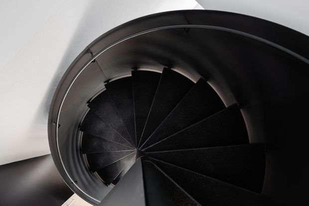 Top-down view of a compact spiral staircase with dark black treads winding around a central point, enclosed by a curved black guardrail and set against white walls. The image emphasizes the geometric, sculptural form and strong light-dark contrast.