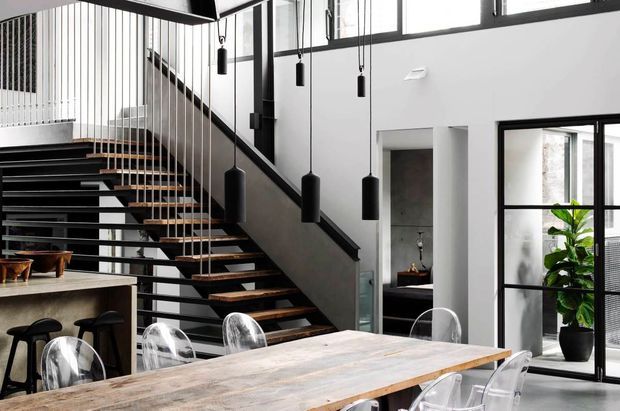 Contemporary open-plan interior centred on a dramatic black steel staircase with timber treads, suspended black pendant lights, and a large timber dining table surrounded by transparent chairs. White walls, black-framed glazing, and a planted area create a bright, graphic, loft-like space.