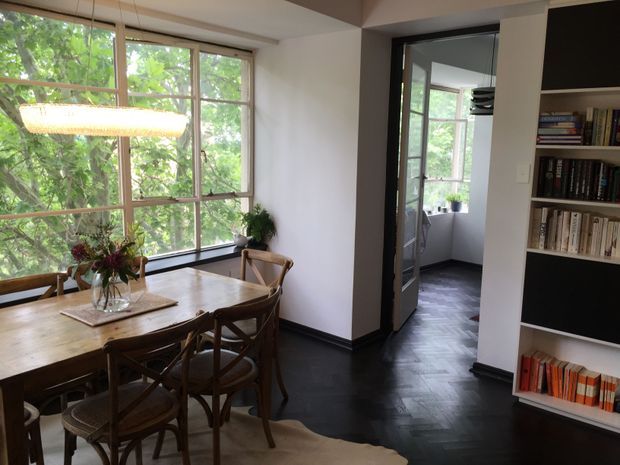 A warm, characterful dining area with a large timber dining table and bentwood-style chairs positioned beside expansive timber-framed windows overlooking leafy greenery. A woven pendant light hangs above the table, while built-in open shelving filled with books occupies the right side. A dark timber herringbone floor runs through the dining room and into an adjoining glazed or glass-door space, with plants and a small seating area visible beyond.