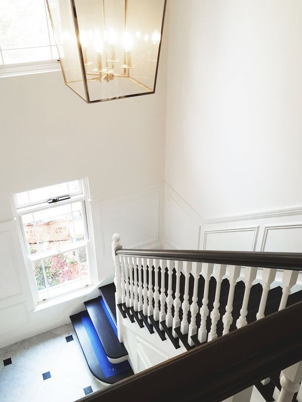 Bright, elegant stairway viewed from an upper landing, featuring a dark timber handrail and stair treads, white turned balusters, white decorative wall panelling, a large window, pale tiled flooring with small dark insets, and a suspended glass-and-brass lantern chandelier. The space has a traditional period-inspired character with abundant natural light.