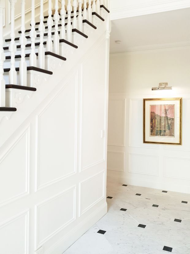 Bright traditional entry hall with a prominent staircase, white painted decorative wall panelling, turned balusters, dark timber stair treads, and a pale stone or marble floor inset with small black square accents. The adjoining hall features matching wainscoting and framed artwork.