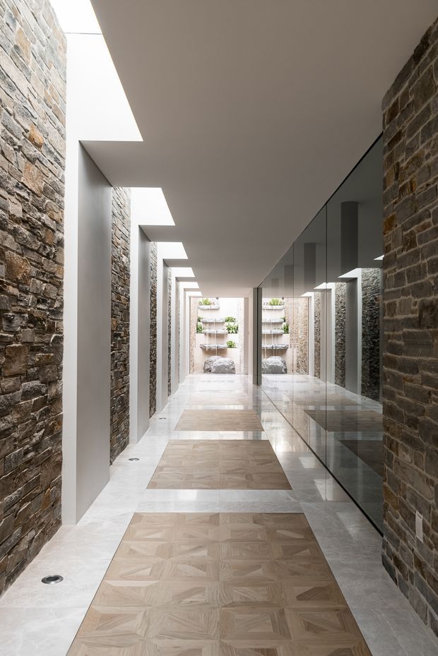 A long, narrow architectural corridor with exposed rough stone walls on both sides, a pale smooth ceiling with repeated skylight openings, and a reflective full-height glass wall along the right. The floor combines large pale stone slabs with inset light timber parquet panels, leading toward a bright courtyard-like end with built-in planters and a water feature. The composition is minimalist and highly geometric, emphasizing natural stone, daylight, repetition, and reflection.