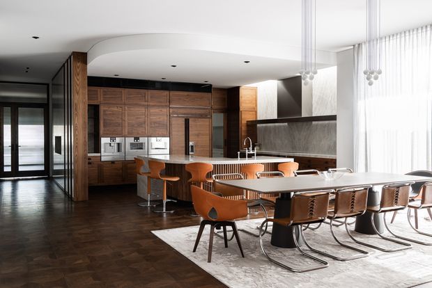 Open-plan contemporary kitchen and dining area with dark herringbone timber flooring, extensive dark timber cabinetry, a large island with integrated appliances and seating, and a long dining table with leather and timber chairs. Full-height sheer curtains provide abundant daylight, while sculptural pendant lights hang above the dining zone. A dark-framed glazed entry or hallway is visible at left.