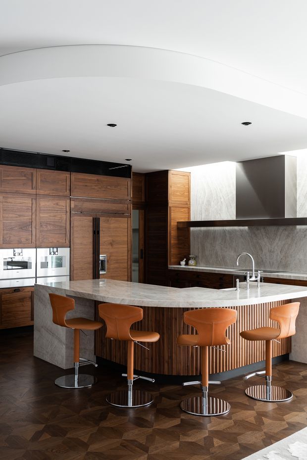 A sophisticated contemporary kitchen with a large curved island, dark walnut cabinetry, vertically fluted timber detailing, and a pale stone benchtop. Four leather bar stools line the island, while integrated appliances and a matching stone splashback sit against the rear wall. The room has a sculptural curved white ceiling with recessed downlights and a dark geometric timber parquet floor.