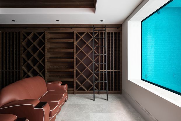 A sophisticated dark-toned media room or home cinema with a large wall-mounted projection screen, built-in dark timber shelving and geometric wine-style display racks, recessed ceiling lighting, and a sculptural brown leather sectional sofa. Pale stone flooring and light walls contrast with the rich timber joinery and leather seating.