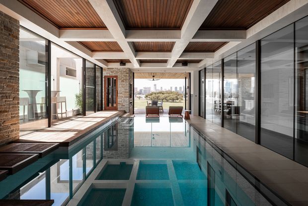 A refined indoor swimming pool extending through a bright, open-plan residential space. The pool is bordered by pale tiled flooring, exposed brick walls, extensive black-framed glazing, and timber-lined ceiling panels set within a bold white concrete beam grid. Lounge chairs sit at the far end, with a city skyline and landscaped outlook visible beyond the glazing. The composition emphasises strong indoor-outdoor connection, reflected light, and a calm contemporary architectural character.