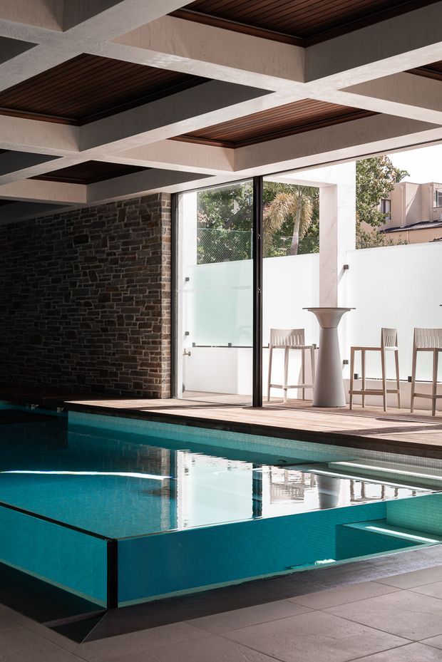 A sophisticated indoor swimming pool area with a turquoise tiled pool, integrated spa or stepped shallow section, and dark stone feature wall. The space opens to a bright terrace through large black-framed glazed doors, with a small bar-height table and timber bar stools. A striking exposed concrete ceiling grid incorporates recessed dark timber slatted panels, creating a geometric architectural feature.