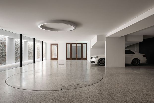 A spacious, highly polished residential garage with a broad open parking and manoeuvring area. The floor is finished in light grey speckled terrazzo or polished concrete, with a large circular inlaid detail. Full-height glazed doors and windows line one side, while white structural columns and walls create a clean, minimalist interior. Two white cars are parked beneath a darker recessed area at right. A large circular ceiling light adds a sculptural feature.