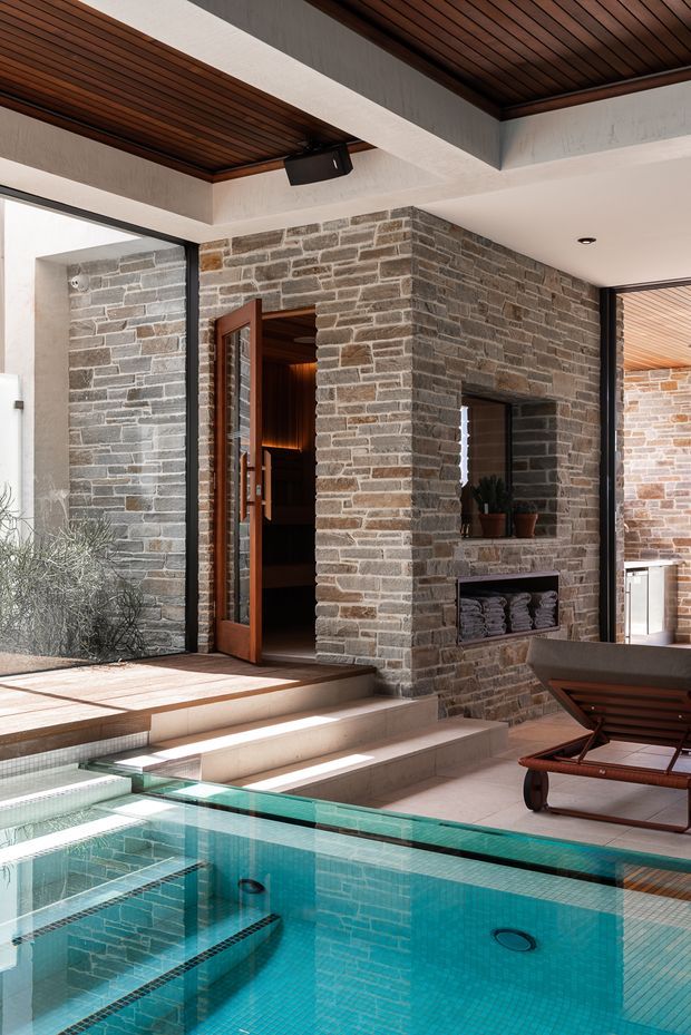 A refined indoor-outdoor pool area centered on a turquoise tiled swimming pool, with broad pale stone steps descending into the water. The space features exposed stacked-stone walls, warm timber slatted ceilings, large black-framed glazed openings, and a timber-framed door. A built-in fireplace or niche with folded towels and potted plants sits within the stone wall, while a dark timber sun lounger occupies the foreground. The architecture combines strong natural materials with abundant daylight and a calm resort-like atmosphere.
