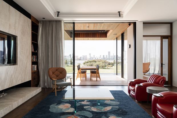 A refined contemporary living room opening onto a covered outdoor terrace through expansive floor-to-ceiling glazing. The room features a large fireplace wall with textured stone or concrete-like cladding, dark timber built-in shelving, a recessed coffered ceiling, dark timber flooring, sculptural patterned rug, glass coffee table, and red leather lounge chairs. The terrace includes a timber dining table and chairs, with an urban skyline visible beyond.