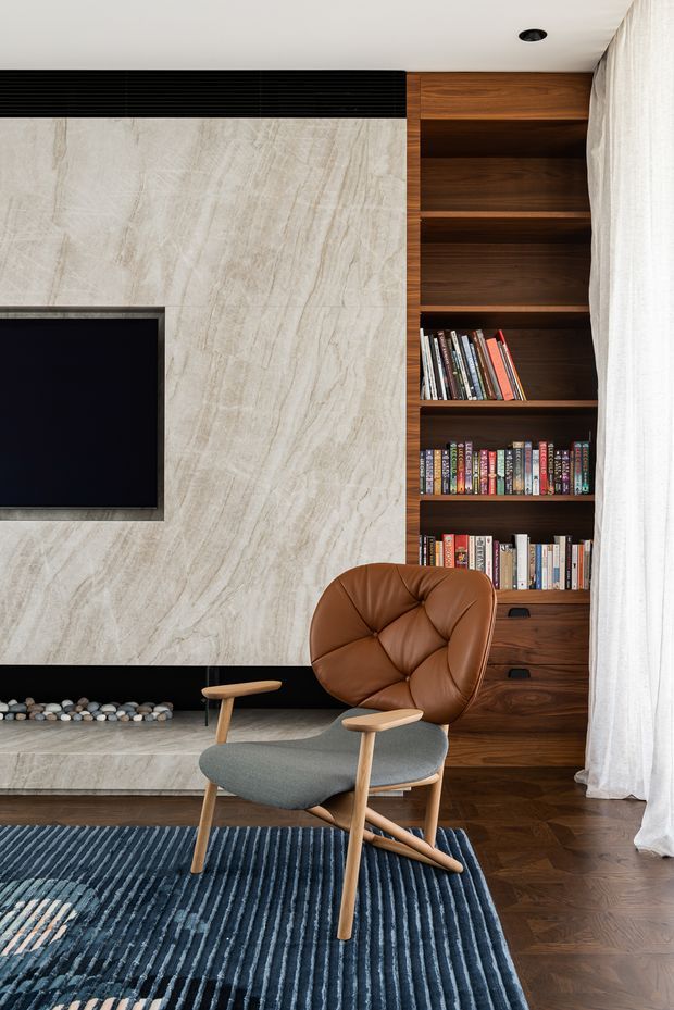 A refined living room featuring a built-in dark timber bookshelf and drawers, a large pale veined stone fireplace wall with a recessed television, and a sculptural mid-century-style lounge chair in brown leather and grey upholstery. Dark herringbone timber flooring, a blue textured rug, sheer white curtains, and a linear black fireplace surround create a warm, sophisticated interior.