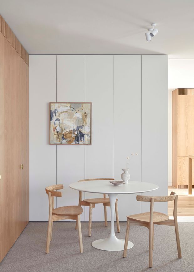 A bright, refined dining area with a round white pedestal table and light timber dining chairs on a soft grey carpet. The room features a full-height pale grey panelled wall, warm timber wall panelling and concealed cabinetry or door surfaces, with minimalist white track lighting and a small abstract artwork. An opening reveals a warm timber-lined adjoining space in the background.